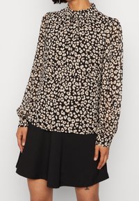 Schwarz-creamfarbige Leopardenmuster-Bluse mit hohem Kragen, langen Ärmeln und gerafftem Detail am Hals. Kombination mit einem schwarzen, ausgestellten Rock.