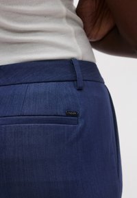 Tmavomodré nohavice z hladkej látky s jedným zadným vreckom a malou čiernou značkou s logom. Úzky strih s jemnou textúrou.