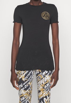 Sort tætsiddende kortærmet skjorte med guld Versace Jeans Couture-logo på brystet, kombineret med sorte, hvide og gyldne mønstrede bukser.