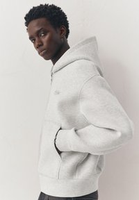 Jeune homme aux cheveux courts et bouclés portant un sweat à capuche gris clair zippé, les mains dans les poches, debout contre un mur uni.