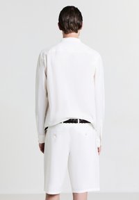 Camicia bianca a trama con maniche lunghe, vista da dietro. Abbinata a pantaloni corti bianchi e una cintura nera intrecciata. Design semplice e pulito.