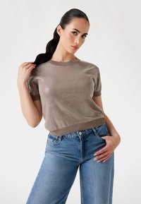 Krátký rukáv, žebrovaný pletený svetr v metalické béžové barvě, s kulatým výstřihem a volným střihem, nošený s modrými džíny s vysokým pasem.