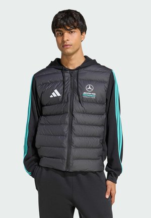 Joven vestido con un chaleco acolchado negro de Mercedes AMG Petronas F1 sobre una sudadera con capucha negra con franjas turquesa, de pie con las manos en los bolsillos.