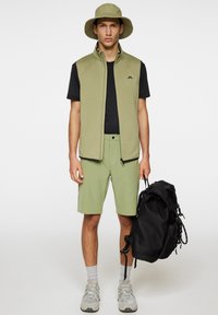 Lättviktig olivväst med hög krage över en svart T-shirt. Olivfärgade shorts, grå strumpor och beige sneakers, bärande en svart ryggsäck.