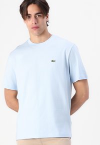 Tricou din bumbac de culoare albastru deschis, cu mâneci scurte, guler rotund și logo verde brodat pe piept. Design simplu, clasic.