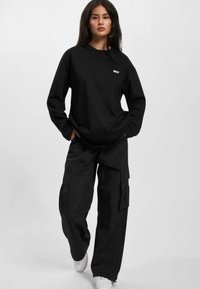 Black long-sleeve shirt with "DEF" logo, paired with loose black cargo pants featuring pockets. Model stands, hands in pockets, wearing white sneakers.