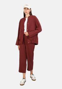 Veste légère bordeaux avec deux poches avant, associée à un pantalon court assorti. Un t-shirt blanc et des baskets complètent la tenue.