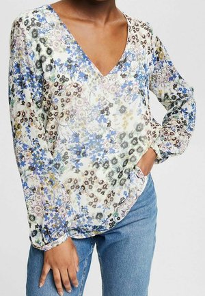 Persona che indossa una camicetta a maniche lunghe con fiori blu, neri e gialli, abbinata a jeans blu, con una mano in tasca.
