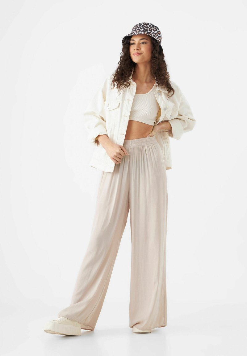 Pantalons larges beiges légers avec une taille smockée, associés à un crop top crème et une veste en denim ivoire. Accessoirisés avec un chapeau seau à imprimé léopard.