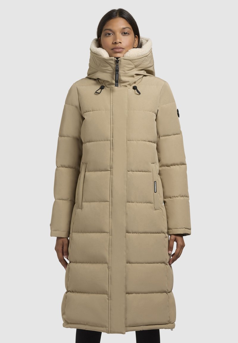 Bežs garš pūkains mētens ar kapuci, ar pāršūtu dizainu, divām sānu kabatām un melnu aparatūru. Mīkstais oderējums redzams apkārt kapucei.