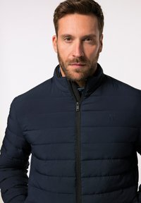 Giacca a vento navy con cuciture orizzontali, collo alto e zip frontale. Presenta un logo discreto sul petto e una superficie liscia.