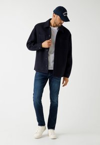 Granatowa koszula-jacket z mieszanki wełny z dużymi przednimi kieszeniami, zapięcie na guziki, zestawiona z białym T-shirtem, ciemnymi dżinsami i białymi trampkami.