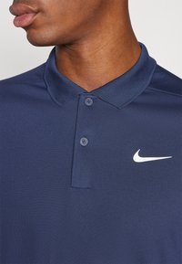 Marinblå polo skjorta gjord av texturerad tyg, med en klassisk krage, två knappar och en vit Nike-logotyp på vänster sida.