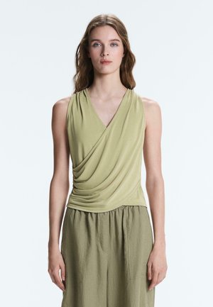 Jonge vrouw met lang haar draagt een mouwloze lichtolijfgroene gedrapeerde top en een losse olijfgroene broek, staand tegen een effen lichte achtergrond.