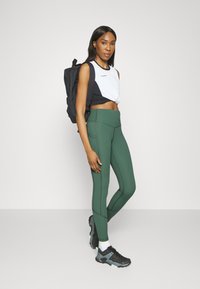 Leggings verts avec une taille haute et des détails de couture, associés à un haut de sport court noir et blanc et un sac à dos noir. Baskets noires avec des accents.