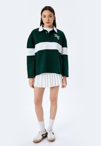 Polo a maniche lunghe verde e bianco a righe con colletto, abbinato a una gonna bianca plissettata e a scarpe da ginnastica bianche con accenti neri.