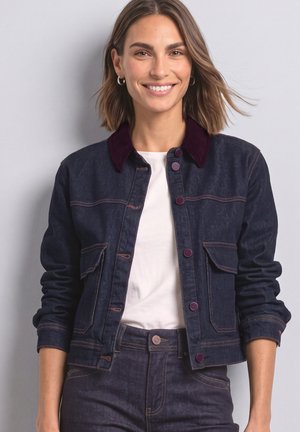 Femme aux cheveux mi-longs souriante, portant une veste en jean foncée avec des boutons bordeaux et un col, une chemise blanche et un jean assorti.