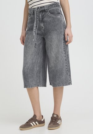 Czarne spodenki dżinsowe o średniej długości ze spranym efektem, z paskiem do wiązania w talii, noszone z brązowymi trampkami Adidas i bluzką w paski.