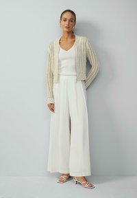 Witte gebreide cardigan met open weefsel, gecombineerd met een witte V-hals top en broek met wijde pijpen. Accessoires omvatten minimalistische sandalen en oorbellen.