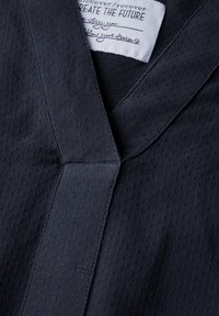 Vêtement bleu marine foncé avec un tissu texturé et perforé, doté d'un col en V et de détails de couture visibles. L'étiquette indique "CRÉER L'AVENIR."