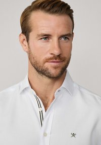 Chemise blanche à manches longues avec un plastron étroit et décoré de rayures vertes. Comprend un petit logo brodé sur la poitrine. Texture lisse.