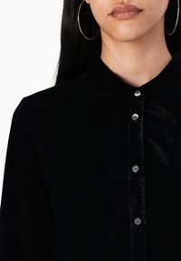 Mustmust särg, millel on krae, hõbedaste nööpidega ja sile tekstuur. Riietus on hästi istuv ja omab õrna läiget.