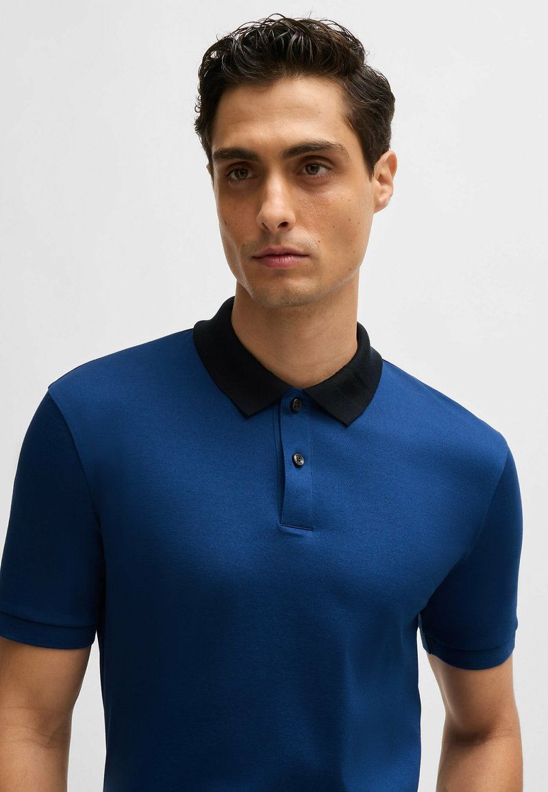 Polo en tissu bleu marine uni avec col noir. Présente une patte de boutonnage à trois boutons et des manches courtes. Texture lisse avec un léger éclat.