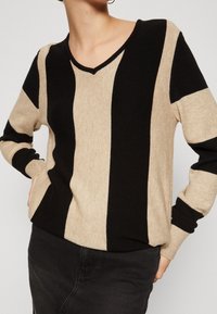 Sweter z dekoltem w serek w naprzemiennie czarne i beżowe paski. Wykonany z miękkiego materiału, z prążkowanymi mankietami i luźnym krojem.
