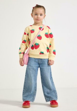 Ung pige iført en gul sweatshirt med jordbærprint, viddebenede jeans, røde sko og en lille lyserød crossbody-taske på hvid baggrund.