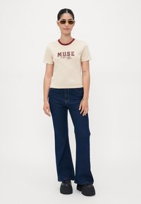 Cream short-sleeve T-shirt with red trim and graphic text "MUSE Energy," paired with dark blue flared jeans and chunky black platform shoes.