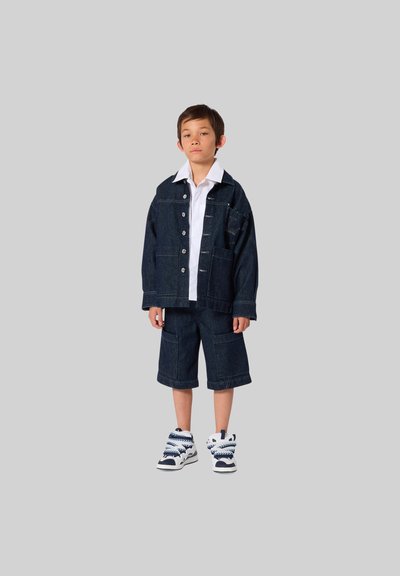 Donkere spijkerjasje met voorsluiting en zakken, gecombineerd met bijpassende shorts. Witte overhemd met kraag eronder en hoge sneakers met blauwe accenten.