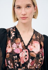 Blomstret kjole med mørk baggrund, prydet med pink og brune blomster, V-udskæring, sorte perleaccentuer og knapdetaljer.