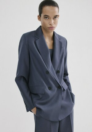 Personne aux cheveux tirés en arrière portant un blazer bleu foncé à double boutonnage et un pantalon assorti, mains dans les poches.