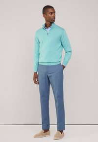 Pullover verde menta con colletto zip sopra una camicia a righe blu e bianche, abbinato a pantaloni azzurri chiari e scarpe casual beige.