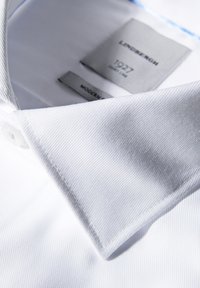 Camicia bianca con una texture a righe, dotata di un colletto rigido e chiusura con bottoni. Le etichette indicano dettagli sul marchio e sul modello.