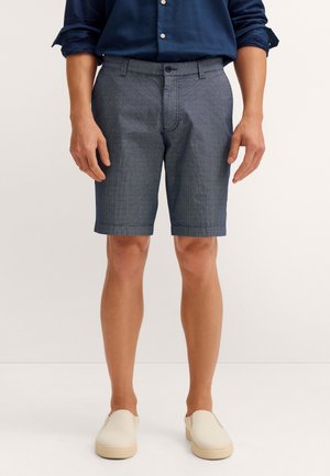 Mann trägt strukturierte graue Shorts, dunkelblaues Langarmhemd mit aufgekrempelten Ärmeln und hellbeige Slipper, steht vor weißem Hintergrund.