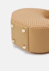 Sac à main beige avec texture tissée, accents en métal doré, clous cubiques et forme arrondie. Panneau latéral en cuir lisse.