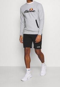 Harmaa huppari, jossa on "ellesse"-logo, mustat yksityiskohdat, sivuvetoketju, yhdistettynä mustiin shortseihin ja valkoisiin urheilu kenkiin. Rentoa aktiivista pukeutumista.