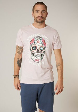 Mann trägt ein hellrosa T-Shirt mit einem bunten floralen Totenkopf und spanischem Text, dazu dunkelblaue Shorts, steht vor einem einfarbigen Hintergrund.