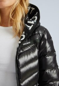 Juodas pūkuotas švarkas su blizgiu paviršiumi, su užtrauktuku ir gobtuvu, vidinė pamušalas su baltu užrašu, ir logotipu.