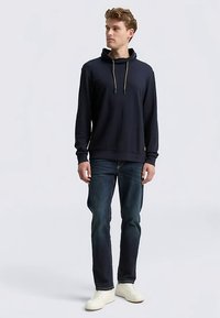 Marineblauer Hoodie mit lockerer Passform, Kordelzug am Hals und gerippten Bündchen, kombiniert mit dunkelblauen Jeans und weißen Turnschuhen.