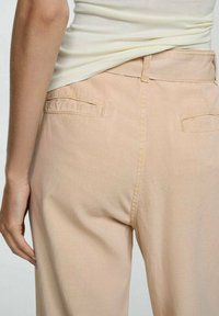 Pantaloni di cotone pesca chiaro con una texture liscia, caratterizzati da due tasche posteriori e dettagli di cucitura discreti lungo la vita e le cuciture.