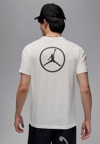 Camiseta crema con un logo negro de Jordan centrado en la parte posterior. Mangas cortas, textura suave y un ajuste casual. Combinada con pantalones cortos oscuros.