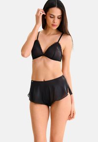 Femme portant un ensemble de lingerie noire transparente avec des bords volants, se tenant avec une main touchant ses cheveux, sur un fond clair uni.