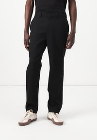 Black tailored trousers with a straight fit, belt loops, and a smooth texture, paired with cream sneakers featuring brown accents.