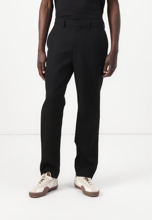 Black tailored trousers with a straight fit, belt loops, and a smooth texture, paired with cream sneakers featuring brown accents.