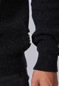 Pull en maille noire avec des poignets côtelés, un tissu texturé et une petite étiquette de marque sur le côté. La couleur est uniforme partout.