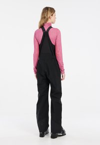 Svarta snöoveraller med justerbara axelband, i en lätt design och lös passform, kombinerade med en figurnära, rosa långärmad topp.