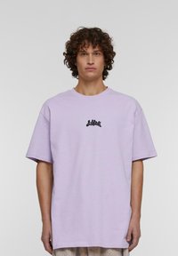 Lys lilla oversized T-skjorte med korte ermer, med en liten svart logo på brystet. Laget av myk, glatt stoff.