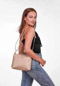 Béžová kožená kabelka přes rameno s hladkým povrchem, štíhlým designem a nastavitelnými popruhy, vystavená na modelce v černém topu a modrých džínách.